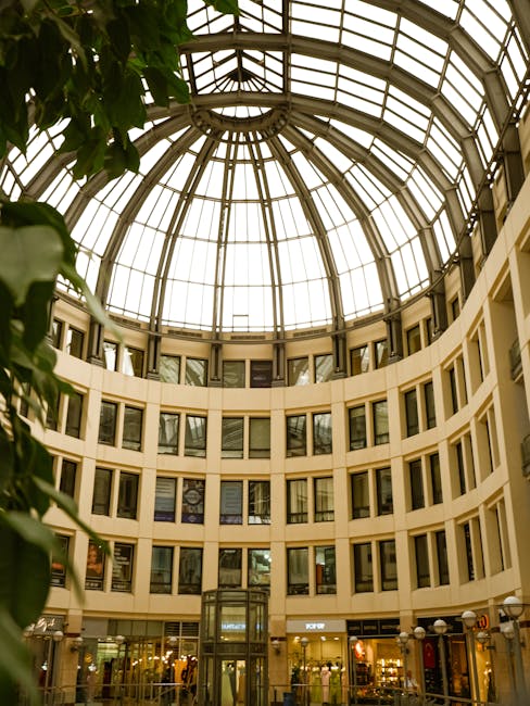 Inside the Bentall Centre shop during a house relocation process, showing an open atrium with a large glass and metal domed roof allowing natural light to illuminate the interior. The circular multi-storey structure features cream-colored walls lined with numerous rectangular windows, some with visible signage or displays. On the ground floor, a retail store with glass storefronts and illuminated displays is visible, alongside a section of the shopping mall interior with people browsing. Furniture, cardboard boxes, and packing materials are being moved by staff from [COMPANY_NAME], who are using trolleys, straps, and blankets to facilitate furniture transport and loading onto a van outside at an accessible loading area. Visible details include individuals carrying a large piece of furniture wrapped in protective fabric, with other boxes and equipment positioned nearby, indicating an active packing and moving operation consistent with a professional removal service focused on office or shop relocations within Kingston.