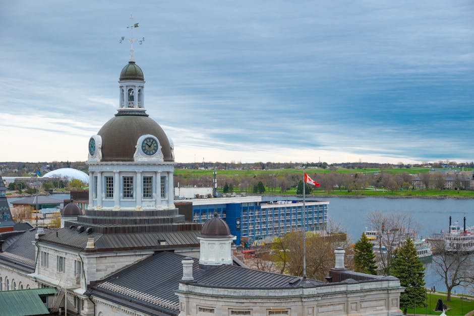 A panoramic view of Kingston with a historic building featuring a large dome and clock faces in the foreground, alongside smaller domed structures and rooftop details. Behind the building, there is a body of water with boats docked along the shoreline, and a bridge connecting the area. In the background, a modern, multi-story blue building is visible, displaying the Canadian flag on a flagpole. The scene is set under a partly cloudy sky, with lush green trees lining the riverbanks and a mix of urban and natural landscape extending into the distance. This image captures the urban environment surrounding the Bentall Centre shop relocation in Kingston, highlighting elements relevant to house removals and furniture transport logistics within a cityscape context, as managed by Man with Van Kingston.