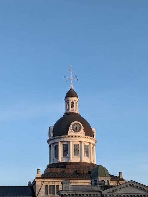 A clear blue sky serves as a backdrop for the historic clock tower of a classical-style building, featuring a rounded black dome with a weather vane on top. The tower has a white base with multiple rectangular windows, some with decorative surrounds, and a prominent clock face below the dome. The building's rooftop area includes smaller structures, such as a green-domed section and ornate stonework cornices. This architectural feature is visible in an urban setting, and the image is relevant to house removals and furniture transport services, such as those provided by Man with Van Kingston, during the process of moving or relocating within Kingston, as seen in the context of planning or executing a home relocation at Canbury Gardens after market day.