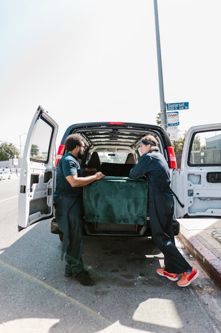 Two movers from Man with Van Kingston are loading a large, dark green upholstered piece of furniture into the back of a white delivery van, which has its rear doors open. They are using their hands to lift and position the item carefully, with one person on each side, working on a paved street amid an outdoor setting with a clear sky. The van is parked next to a curb, and visible elements include cardboard boxes, bubble wrap, and packing blankets that suggest a home relocation or furniture transport process. The environment features street signage, utility poles, and some trees in the background, typical of a residential or commercial area in Kingston. The scene captures a professional move involving careful handling of household furniture as part of a packing and loading process at a moving site, with Man with Van Kingston conducting the home removal task efficiently.