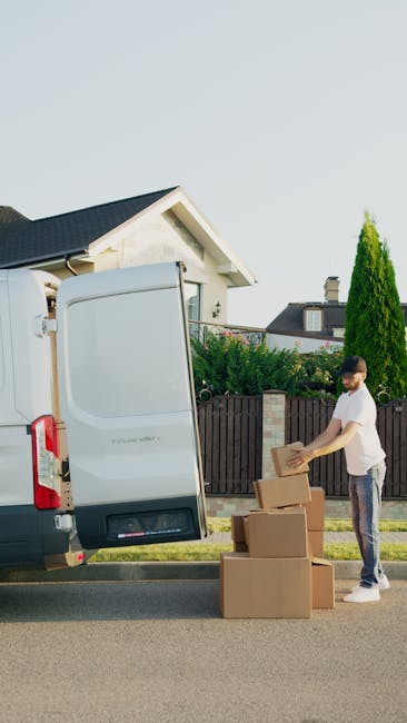 A person wearing a white t-shirt and jeans is seen standing on the pavement beside a large, white moving van with its side door open, engaged in loading or unloading cardboard boxes. The van is parked outside a residential property with a small front garden, a wooden fence, and tall green bushes. Inside the van, visible packing materials include plastic wrap and cardboard boxes. The individual is handling a box, with several other boxes stacked on the pavement nearby, ready for moving or home relocation purposes. The scene is set during daylight with clear weather, emphasizing the physical process of furniture transport and packing associated with professional removals by companies like Man with Van Kingston.