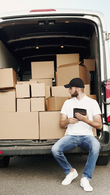 Two movers from Man with Van Kingston are loading a large, dark green upholstered piece of furniture into the back of a white delivery van, which has its rear doors open. They are using their hands to lift and position the item carefully, with one person on each side, working on a paved street amid an outdoor setting with a clear sky. The van is parked next to a curb, and visible elements include cardboard boxes, bubble wrap, and packing blankets that suggest a home relocation or furniture transport process. The environment features street signage, utility poles, and some trees in the background, typical of a residential or commercial area in Kingston. The scene captures a professional move involving careful handling of household furniture as part of a packing and loading process at a moving site, with Man with Van Kingston conducting the home removal task efficiently.