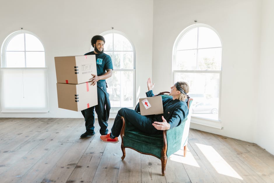 A man and a woman inside a well-lit room during a home relocation, both smiling and holding packing items. The man, wearing a maroon t-shirt and orange pants, holds a small cardboard box labeled 'Frag