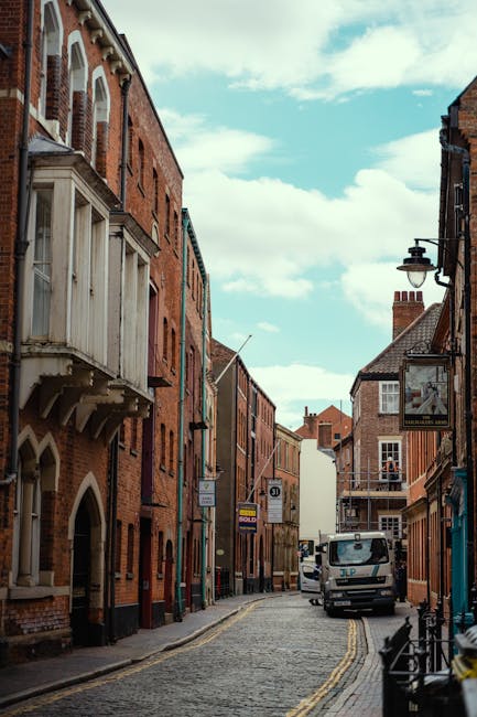 A narrow cobblestone street in a residential area with red-brick terraced houses on both sides. Some buildings feature small bay windows and decorative brickwork, while others have wooden balconies and traditional street lamps attached to the facades. A white van with the logo of a removals company is parked on the street, adjacent to the pavement, partly blocking the curb. Behind the van, there are a few cardboard boxes and wrapped furniture items being loaded or unloaded, suggesting a home relocation process. The sky above is partly cloudy with patches of blue visible, and the street is quiet with no pedestrians. The scene reflects a typical setting for furniture transport and packing during house removals in Kingston, with the van positioned appropriately for parking permit rules for removals as indicated on the Kingston Council road signage.