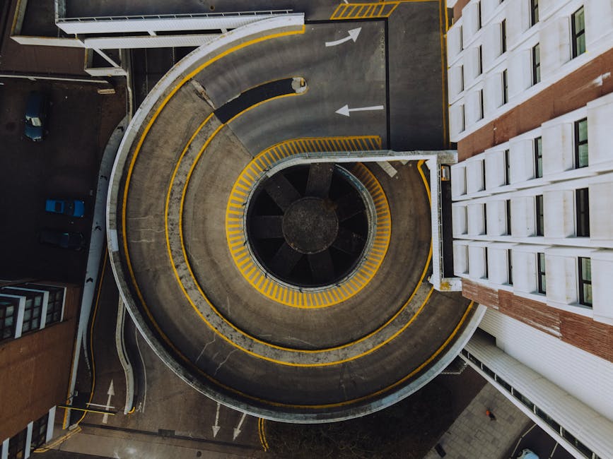 An aerial view of a multi-story, circular parking structure with yellow safety lines and directional arrows painted on the concrete surface, indicating driving lanes and vehicle flow. The parking garage is surrounded by the edges of adjacent buildings, including a white modern apartment block with multiple windows on one side and a darker, brick-finished building on the other. The garage appears empty except for a few parked cars in a small section, and the lighting suggests daytime. The image captures the process of home relocation logistics, as it relates to furniture transportation and loading for a local move, with the parking structure providing space for a van belonging to Man with Van Kingston, situated near the building's entrance, ready for loading or unloading furniture, boxes, and packing materials during a residential move.