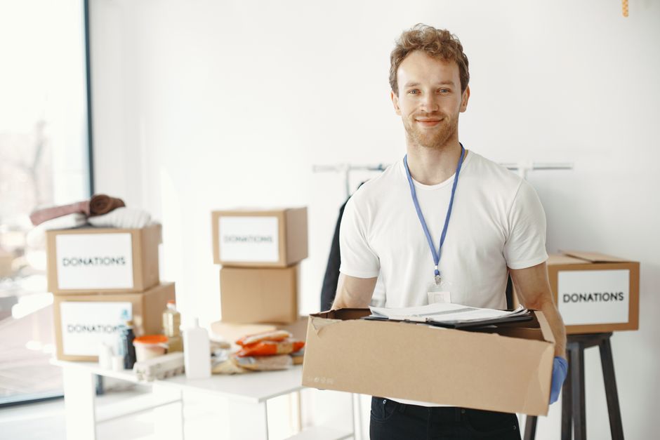 A person wearing a white shirt and a blue disposable glove is seen placing a glass jar into a cardboard donation box labelled 'DONATIONS,' which is set on a table covered with various items including 