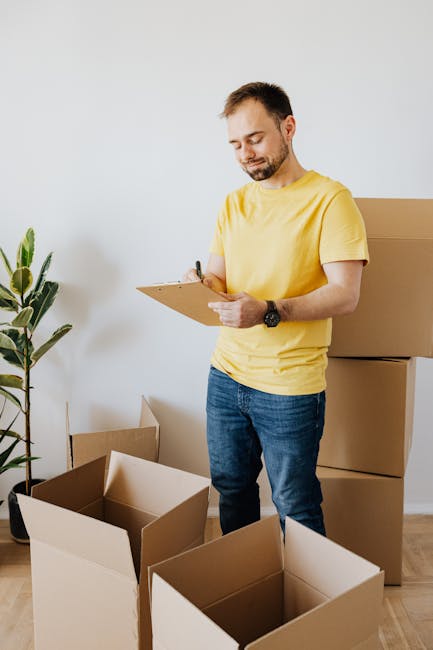A man with a beard and short dark hair, wearing a yellow t-shirt and a black wristwatch, is standing indoors on a wooden floor near a white wall. He is holding a clipboard and a pen, appearing to chec
