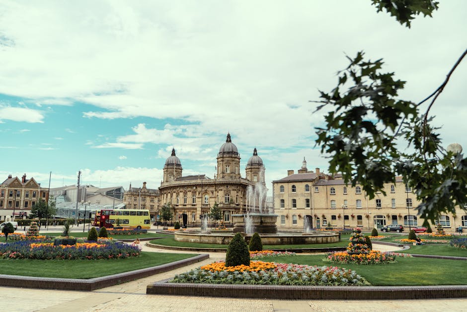 A wide-angle view of a city square in Kingston, KT1, featuring a central ornate stone fountain with water jets, surrounded by well-maintained flower beds and neatly trimmed bushes. In the background, there are historic multi-story buildings with classical architecture, including domed turrets and decorative facades, indicative of a traditional European style. A yellow double-decker bus is visible on the street to the left, moving past the square, while the foreground shows part of a green tree branch with leaves partially framing the scene. The sky above is partly cloudy with patches of blue, and the overall atmosphere suggests a peaceful urban environment. This setting could relate to a home relocation scenario where furniture or boxes might be transported through the busy streets, with [COMPANY_NAME] providing professional removals and packing services in Kingston.