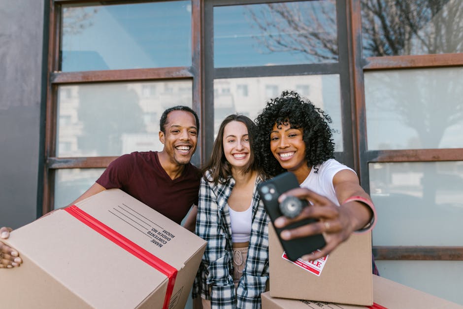 Three individuals standing outside a building with large windows, holding cardboard boxes sealed with red packing tape, as part of a home relocation process. The person on the left is a man wearing a maroon t-shirt, smiling while supporting a box labeled 'HOME MOVING BOXES.' In the middle, a woman with long brown hair dressed in a plaid shirt is smiling broadly. On the right, a woman with curly black hair wearing a white t-shirt is taking a selfie with a smartphone and smiling, while also holding a box. The background shows the building's exterior, with reflections of trees and other buildings visible in the windows. The scene depicts packing, moving logistics, and furniture transport, consistent with house removals services for Kingston University students, as provided by Man with Van Kingston.