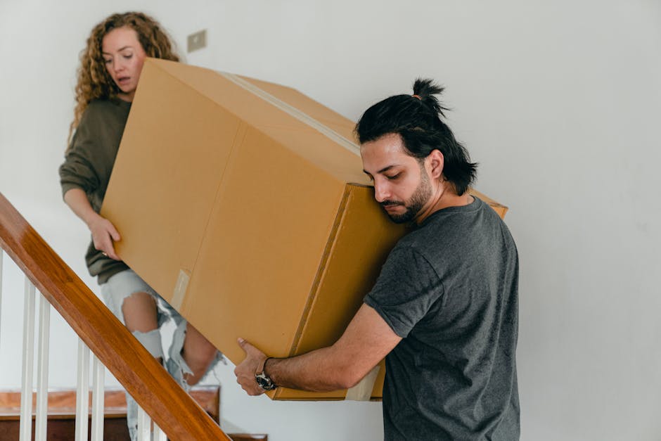 A man with dark hair tied back in a ponytail and wearing a grey t-shirt is lifting a large cardboard box during a home relocation inside a property. He is positioned on a staircase, gripping the box firmly with both hands, and appears focused on the loading process. Behind him, a woman with curly hair, dressed in a brown top and ripped jeans, is standing on the stairs holding another similar cardboard box. The staircase has a wooden handrail and white balustrades, and the surrounding environment includes a plain white wall, suggesting an indoor setting. The boxes are sealed with brown packing tape, indicating careful packing for moving. This scene is part of furniture transport and packing and moving activities executed by a professional service like Man with Van Kingston, supporting house removals and relocation logistics.