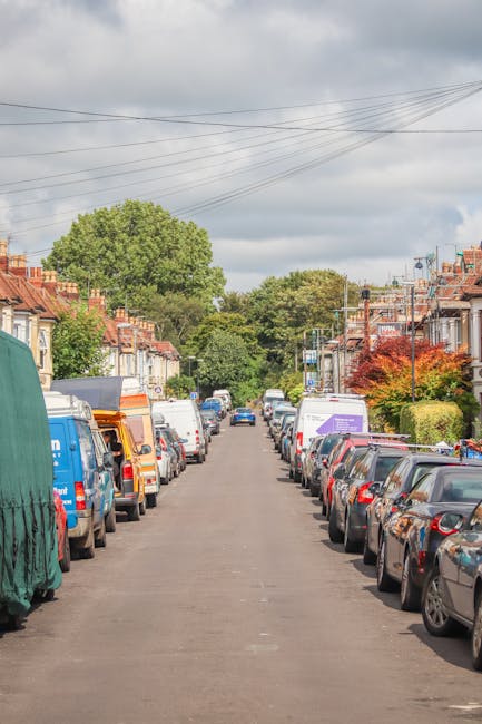 A residential street scene showing a narrow pavement lined with parked cars on both sides, including sedans, hatchbacks, and vans, some of which are covered with protective tarps or fitted with roof racks. At the far end of the street, there are trees with lush green foliage and a few homes with brick facades, pitched roofs, and small front gardens. Overhead, a network of power lines crosses the cloudy sky. In the foreground, part of a white van with visible branding and a loading ramp suggests a home relocation process, with the van positioned close to a house entrance indicating ongoing or upcoming furniture transport. The scene illustrates typical urban moving logistics where careful loading, packing materials, and vehicle suitability are considerations for house removals, with Man with Van Kingston potentially involved in facilitating the furniture transport and packing tasks associated with the near-kingston moving service.
