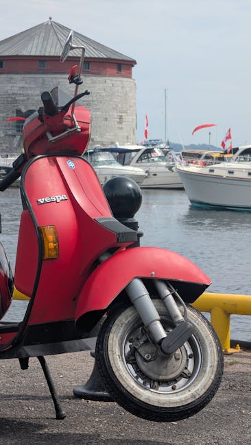 A close-up of a red Vespa scooter parked on a paved surface near a marina, with its front wheel and handlebars visible. The scooter features a black seat, a side mirror, and a small storage compartment on the rear. In the background, there are several boats moored in the water, with white hulls and red flags attached to some of the boats, and a large round stone building with a conical roof. The scene is outdoors with overcast lighting, and the setting appears to be within a harbour area near a waterfront. The image is associated with house removals and moving services, reflecting aspects of transport and logistics involved in property relocation, as demonstrated by the surrounding environment and context.