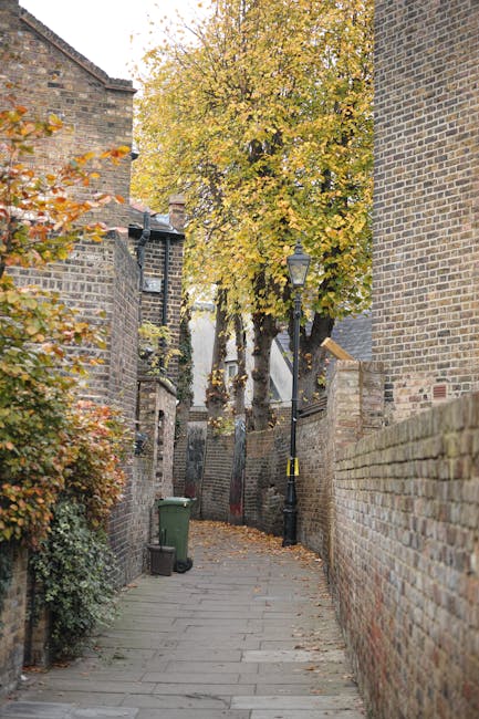 A residential street scene showing a narrow pavement lined with parked cars on both sides, including sedans, hatchbacks, and vans, some of which are covered with protective tarps or fitted with roof racks. At the far end of the street, there are trees with lush green foliage and a few homes with brick facades, pitched roofs, and small front gardens. Overhead, a network of power lines crosses the cloudy sky. In the foreground, part of a white van with visible branding and a loading ramp suggests a home relocation process, with the van positioned close to a house entrance indicating ongoing or upcoming furniture transport. The scene illustrates typical urban moving logistics where careful loading, packing materials, and vehicle suitability are considerations for house removals, with Man with Van Kingston potentially involved in facilitating the furniture transport and packing tasks associated with the near-kingston moving service.